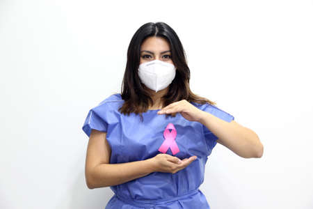 Latin woman with protection mask, disposable blue surgical gown and pink ribbon for campaign against breast cancerの写真素材