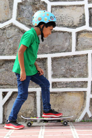 Little dark-haired Latino boy plays in the park with his skateboard wearing a helmet and...