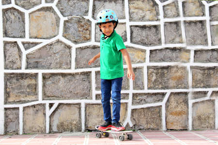 Little dark-haired Latino boy plays in the park with his skateboard wearing a helmet and...