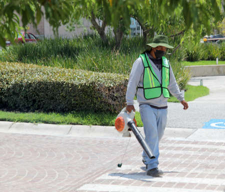 Man wearing a mask, high visibility safety vest and hat works cleaning the streets with a leaf blower in the sunの写真素材