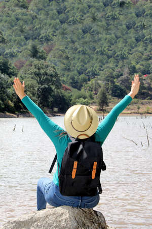 Latin adult woman with hat and sunglasses sitting on a stone next to a lake makes the victory sign...