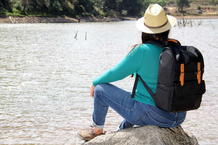 Latin adult woman with hat and sunglasses sitting on a stone next to a lake makes the victory sign happy, accomplished, with achievements and goals achievedの写真素材