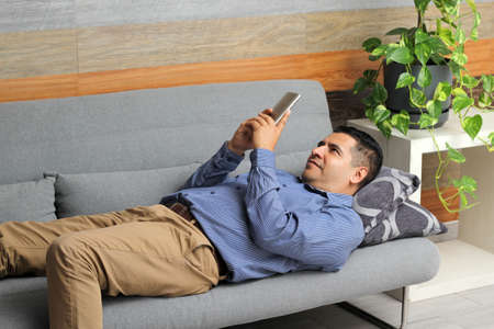 Latin adult man uses his cell phone in an armchair in a room makes calls, video calls, sends messages, buys online, uses social networks and looks for loveの写真素材