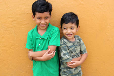 Two dark-haired latin male children standing in front of a yellow wall live in poverty without toys, dream, play, imagine a happy childhoodの写真素材