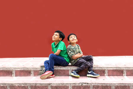 Two dark-haired Latino male children sitting on the ground in a park living in poverty without toys,...