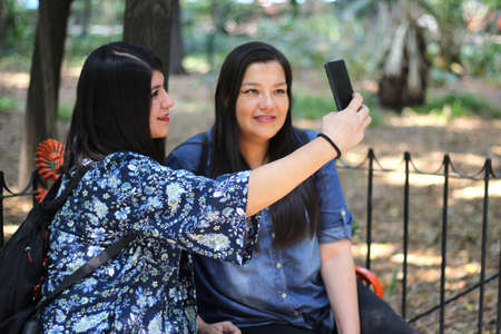 Two Latin women sister friends are sitting on a park bench outdoors using their cell phone to check...