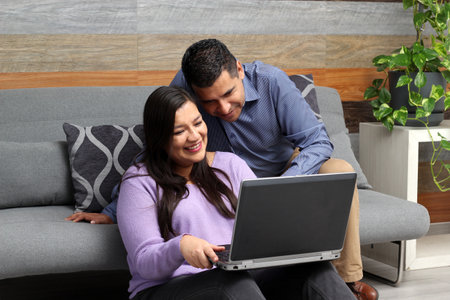 Latino adult man and woman couple use their laptop in the living room to shop online, make payments, plan trips, view photos and make video callsの写真素材