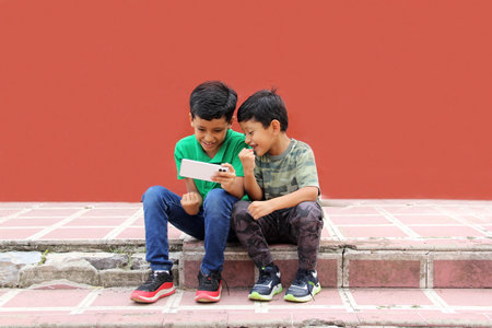 Two dark-haired Latino children who are brothers or friends who live in poverty play video games with a cell phone, make video calls and take selfiesの写真素材