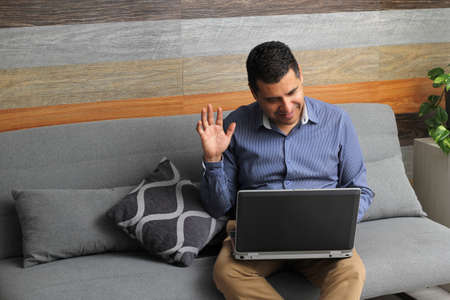 Single Latino adult man does home office in his living room with laptop and cell phone, creates...