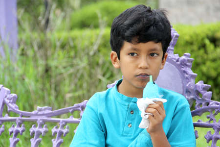 Poor Latino dark-haired boy with a blue t-shirt sitting on a park bench eating an ice pop because of...