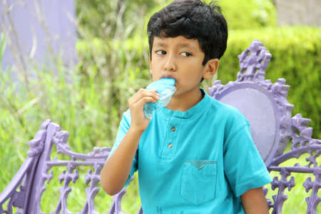 Poor Latino dark-haired boy with a blue t-shirt sitting on a park bench eating an ice pop because of...