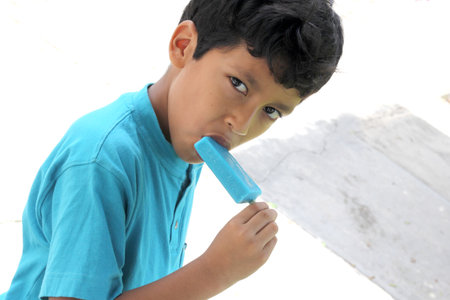 Poor Latino dark-haired boy with a blue t-shirt sitting on a park bench eating an ice pop because of...
