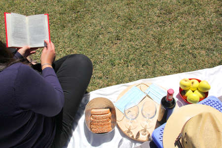latin woman with book and cell phone sitting on the grass with picnic basket, wine and food outdoors in the forestの写真素材