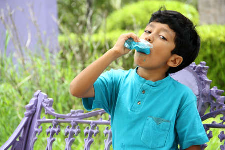 Poor Latino dark-haired boy with a blue t-shirt sitting on a park bench eating an ice pop because of the heat wave and cooling off in poverty by affecting his teeth with sweets andの写真素材