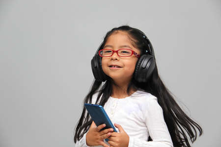 4-year-old brunette Latin girl with glasses listens to music in her headphones connected to her cell phone dances, sings and enjoys the rhythm happy and excitedの写真素材