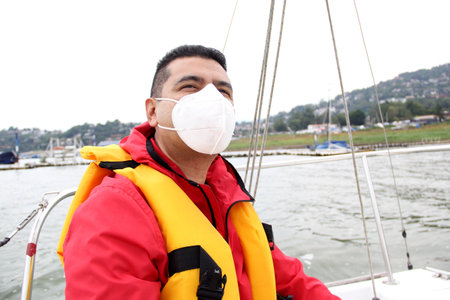 Latino adult man drives a sailboat on the lake with windbreaker and life jacket, wears face masks in...