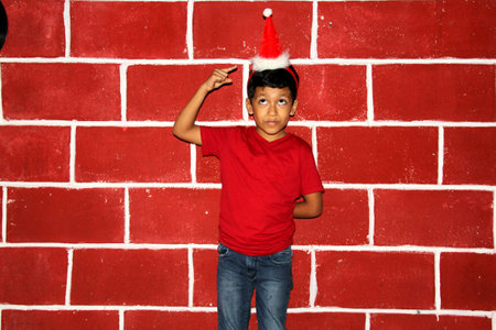 Latin brown boy very happy with the arrival of Christmas wears a red hat in front of a brick wallの写真素材