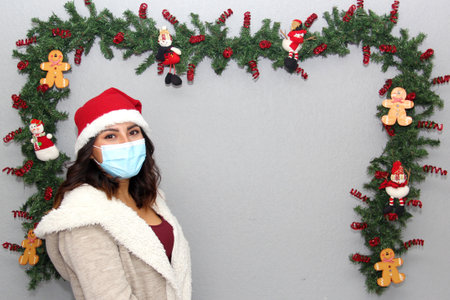 latin woman with protection mask, sweater and christmas hat on gray background with christmas decoration, new normalの写真素材