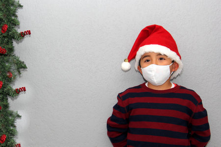7-year-old latin boy with protection mask, sweater and Christmas hat on gray background with Christmas decoration, new normal covid-19の写真素材