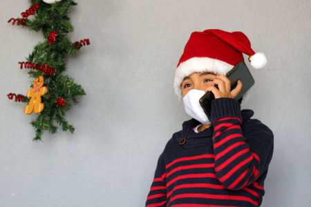 Latin boy with protection mask and Santa Claus hat talking with mobile video call and Christmas decoration, new normal covid-19の写真素材