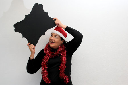 Latin adult woman with hat and Christmas garland shows banner and blank notebook to put a message of...