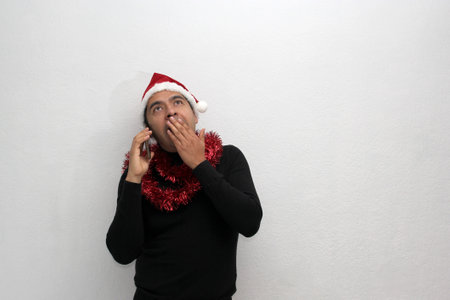 Dark-haired Latin adult man with hat and Christmas garland shows his enthusiasm and happiness for the arrival of December and celebrating Christmasの写真素材