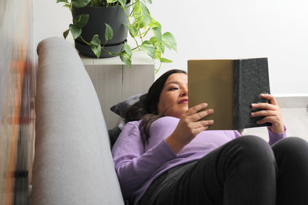 Latin adult woman lying on a sofa in the living room reads a book relaxed, enjoys her hobby in her...
