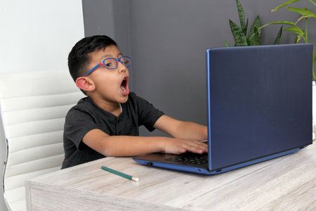 7-year-old Latino boy with glasses does home schooling takes online classes at home on a desk with a...
