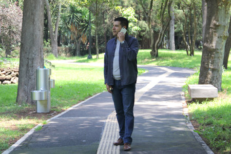 Dark-haired 40-year-old Latino man in a park uses his cell phone to work remotely, make calls, date, shop online while taking a walkの写真素材
