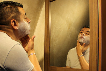 Dark-haired 40-year-old Latino man does his beauty routine, shaves with shaving cream and soap to avoid folliculitisの写真素材