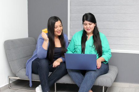 Two friendly Latina women use their laptop and credit card to make purchases online are surprised by...