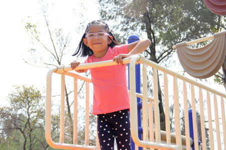 4 year old Latina brunette girl with glasses plays on the playground slide in the park enjoys the...
