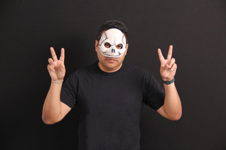 Mexican adult man wears skull mask and makes victory sign with fingers to celebrate Day of the Dead...