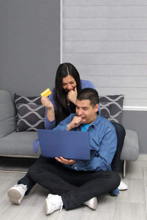 Couple of 40 years old dark-skinned Latino man and woman use a laptop and a credit card to make...