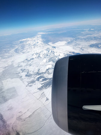 Window view of airplane flying over snowy mountains in winter time with cold, snow and freezing
