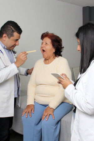 80-year-old woman has her eyes, ears, and throat examined by a dark-skinned Latino doctor and his intern in the office as a checkupのeditorial素材