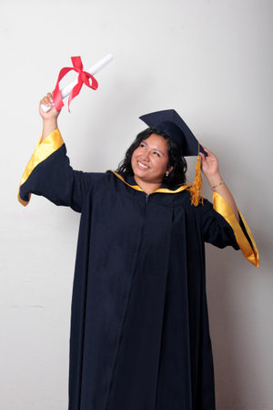 Young brunette Latina woman dressed in a cap and gown shows her diploma proudly, satisfied and happy...