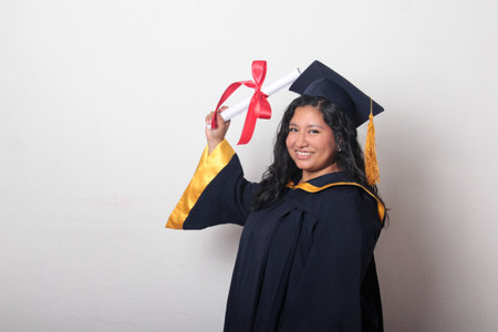 Young brunette Latina woman dressed in a cap and gown shows her diploma proudly, satisfied and happy to finish her career at schoolの写真素材