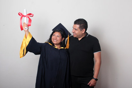 Proud and satisfied single dad with his brunette Latina daughter, a graduate dressed in a cap and...