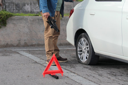 Dark-skinned Latino adult man uses a hydraulic jack to change his car's flat tire on the road