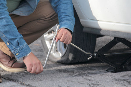 Dark-skinned Latino adult man uses a hydraulic jack to change his car's flat tire on the roadの写真素材