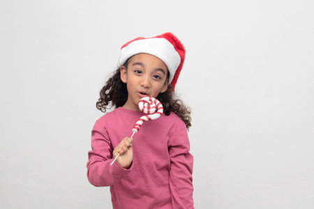 4-year-old Latina girl with curly brown hair wearing a santa hat sings with a candy cane to celebrate Christmas and New Yearの写真素材