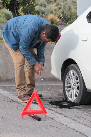 Adult Latino man changes a flat tire with a lug wrench and removes the lug nuts to put on the spare...