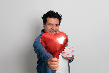 40-year-old Latino man with dark hair and curly hair celebrates Valentine's Day and shows his love with a heart-shaped balloon, glasses, a gift, and a teddy bearの写真素材