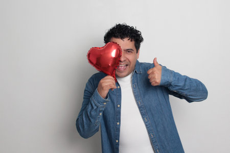 40-year-old Latino man with dark hair and curly hair celebrates Valentine's Day and shows his love with a heart-shaped balloon, glasses, a gift, and a teddy bearの写真素材