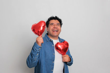 40-year-old Latino man with dark hair and curly hair celebrates Valentine's Day and shows his love with a heart-shaped balloon, glasses, a gift, and a teddy bearの写真素材