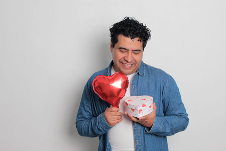 40-year-old Latino man with dark hair and curly hair celebrates Valentine's Day and shows his love with a heart-shaped balloon, glasses, a gift, and a teddy bearの写真素材