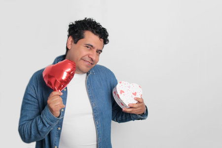 40-year-old Latino man with dark hair and curly hair celebrates Valentine's Day and shows his love with a heart-shaped balloon, glasses, a gift, and a teddy bearの写真素材