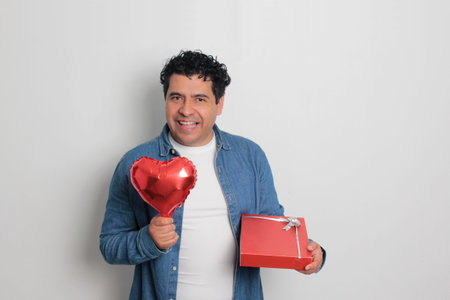 40-year-old Latino man with dark hair and curly hair celebrates Valentine's Day and shows his love with a heart-shaped balloon, glasses, a gift, and a teddy bearの写真素材