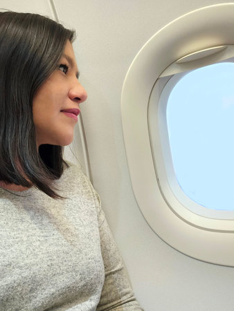 Adult Latina woman looks out the airplane window at the blue sky landscape while sitting alone in...